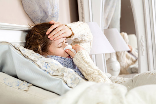 Flu. Closeup image of frustrated sick woman lying in bed in thick blue scarf holding tissue by her nose and touching her head with closed eyes.