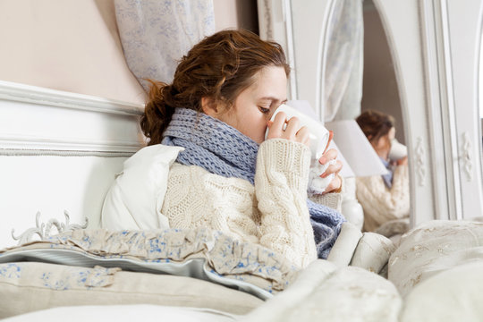 Sick woman with cup of tea. Closeup image of young frustrated woman in knitted blue scarf holding and drinking a cup of tea while sitting in bed of her room.