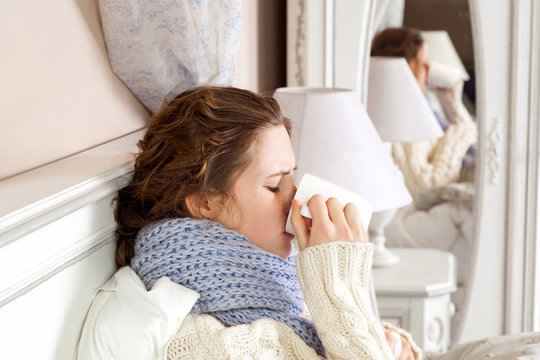Sick woman with cup of tea. Closeup image of young frustrated woman in knitted blue scarf holding and drinking a cup of tea while sitting in bed of her room.