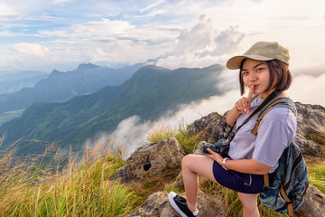 Happy hiker asian cute teens girl with dslr camera backpack and caps standing poses finger up touch the lips on top of mountain at viewpoint Phu Chi Fa Forest Park in winter, Chiang Rai, Thailand