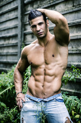 Muscular young latino man shirtless in jeans in front of concrete wall, showing sexy body with ripped abs, pecs and arms