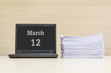 Closeup computer laptop with march 12 word on the center of screen in calendar concept and pile of work paper on wood desk and wood wall in work room textured background with copy space