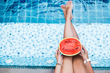 A girl holds half a red watermelon stretching long, tanned legs over a blue pool, relaxing on a tropical island in a hotel, eating healthy, fruit diet, summer style