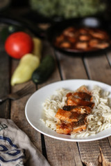 Fish in tomato sauce and pasta in a white plate on a wooden surface (on a table) and vegetables