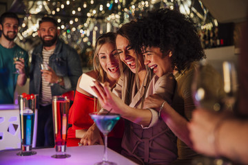 Three women are taking selfies together on a smartphone while enjoying drinks in a night club.