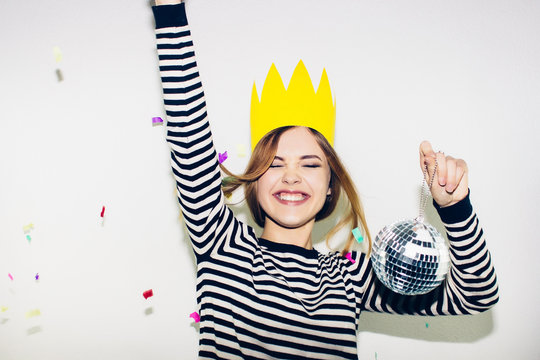 Birthday party, new year carnival. Young smiling woman on white background celebrating brightful event, wears stripped dress and yellow crown. Sparkling confetti, having fun, dancing, laugh, smile.