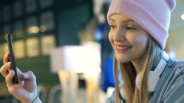 Pretty, blonde girl in pink beanie is doing selfies on smartphone in the cafe

