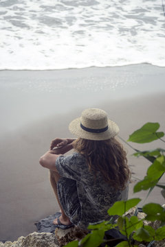 Indonesia, Java, woman wearing straw hat sitting on rock on the beach looking to the sea