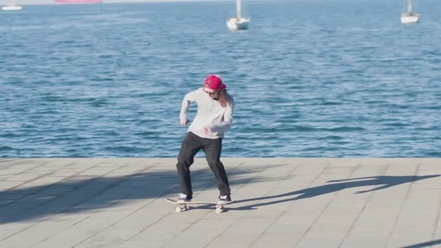 CLOSE UP SLOW MOTION: Young pro skateboarder skateboarding and jumping ollie flip trick on promenade along the coast on sunny summer day with beautiful green cape and the ocean in the background