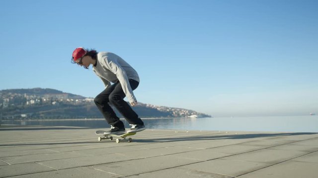 CLOSE UP SLOW MOTION: Young pro skateboarder skateboarding and jumping ollie flip trick on promenade along the coast in sunny summer. Skateboarder jumping kickflip trick with skateboard near the ocean