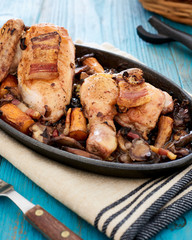 Baked chicken in beer sauce with mushroom, carrots and bacon in an iron cast baking pan on a blue table, with some bread slices in a basket