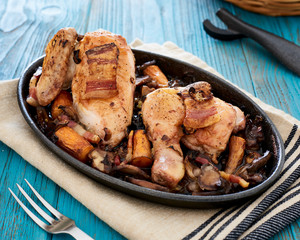 Baked chicken in beer sauce with mushroom, carrots and bacon in an iron cast baking pan on a blue table, with some bread slices in a basket