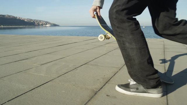 SLOW MOTION CLOSE UP: Unrecognizable skateboarder walks on concrete shore, jumps on skateboard and starts skating. Skater jumping on skate and starts cruising on seaside pavement on sunny day