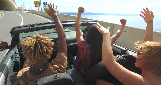 Back view of friends waving arms driving on the coastal road in convertible