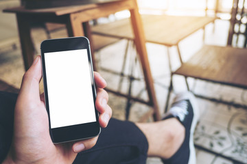 Mockup image of hand holding black mobile phone with blank white screen on thigh with black canvas shoes at vintage tile floor in cafe 