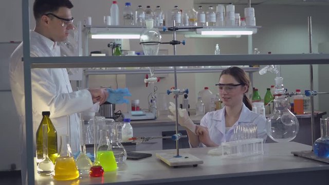 Happy smiling scientists preparing to start working. Two people talking wearing sterile blue gloves. Attractive young caucasian woman sitting at the working place desk with different medical lab