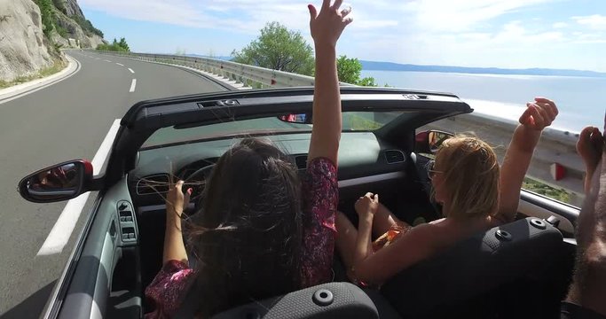 Two attractive girls waving their arms driving in convertible car with friends