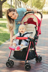 Young mother talking to smiling baby in pink stroller. Parents walking outdoors with child in summer pram.