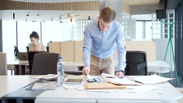 young working entrepreneur look for information among piles of papers, documents and folder for business analytics tasks staying at the table in the office open space during sunny day