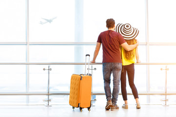 Young couple in colorful t-shirts waiting with baggage near the window at the departure area of the airport during their summer vacation