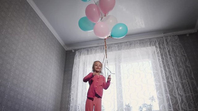 Happy little girl standing near the window on the bed in the nursery with the bunch of air balloons above. Small female child is jumping up on the sofa in the room trying to catch colourful balls.
