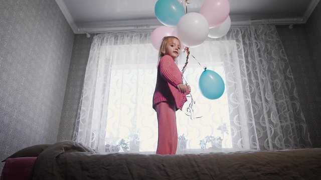 Happy little girl jumping near the window on the bed in the nursery with the bunch of air balloons in her hands and looking at the camera. Small female child is happily bouncing on the sofa.