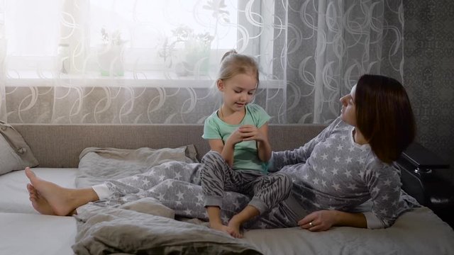 Happy loving family is spending time together in bedtime playing and hugging. Smiling daughter and mother dressed in pyjamas are lying in bed together. Young mom is touching gently her child.