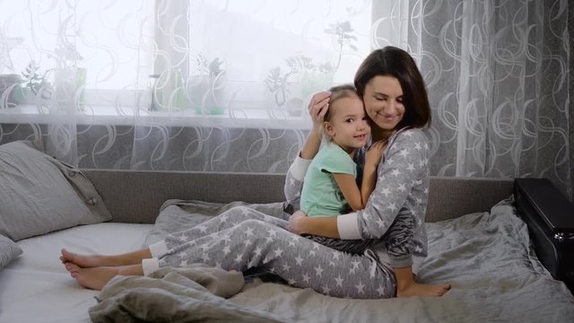 Young mother, dressed in pajamas and her daughter having fun on the bed in the bedroom, enjoying each other's company. Girl tries to kiss his mother, the two beauties having fun and laughing