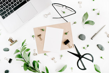 White office desk workspace with paper blank, green leaves and office supplies. Laptop, scissors, glasses on white background. Flat lay, top view, mockup.