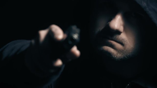 Hooded Man Holds Gun Angry Rack Focus. a hooded man holds a gun up and to the side with an angry tone. Camera racks focus to tip of the gun
