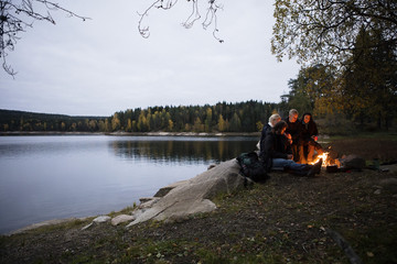 Young Friends Sitting Near Bonfire By Lake At Dusk