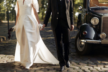 luxury elegant wedding couple walking and holding hands close up at stylish black car in light in...