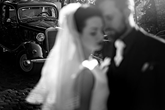 gorgeous elegant bride and stylish groom embracing, gentle touch in light. unusual luxury wedding in retro style. focus on car. unusual view. black white photo
