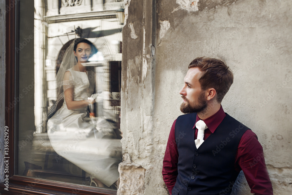 Wall mural elegant gorgeous bride drinking coffee in window and stylish groom posing at building in street outd