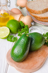 Fresh, green avocado, lime, parsley, sea salt, pepper, bread and eggs for sandwich on a white wooden background