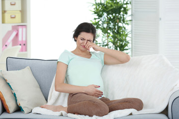 Young pregnant woman crying while sitting on sofa at home