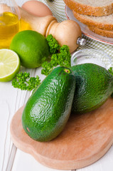 Fresh, green avocado, lime, parsley, sea salt, pepper, bread and eggs for sandwich on a white wooden background