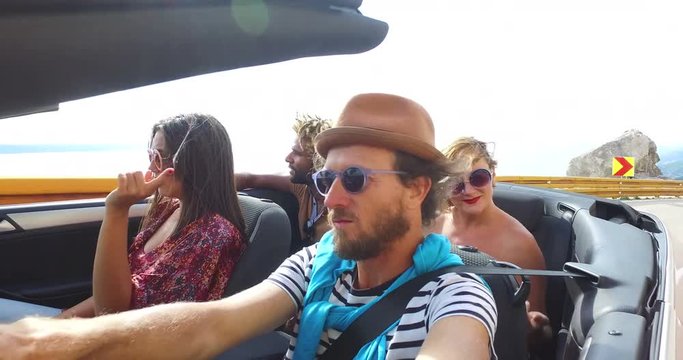 Bearded hipster man driving in convertible on winding motorway with friends