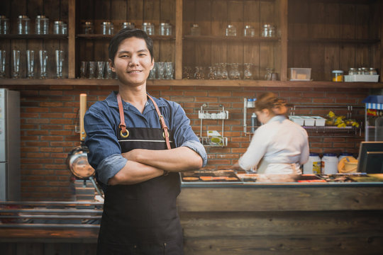 Successful small business owner standing with crossed arms with employee in background preparing coffee. Portrait of asian young male cafe owner .