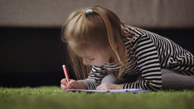 Girl with blond hair, dressed in a gray striped dress and tights is sitting on the green carpet and rasskrashivaet pictures in an album for drawing. She likes to paint markers