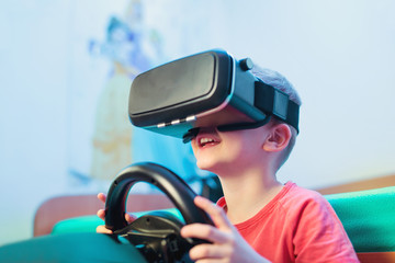 Happy little boy in virtual reality glasses playing video game with racing wheels at home