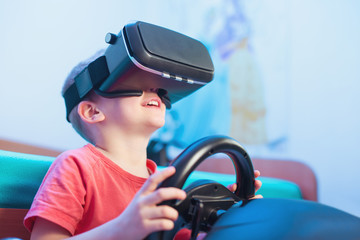 Happy little boy in virtual reality glasses playing video game with racing wheels at home