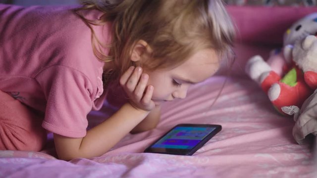 Beautiful child with blond hair, in a pink shirt lying on crib and looking cartoons on her mum mobile phone. Girl keen picture, so not distracted no matter what happens