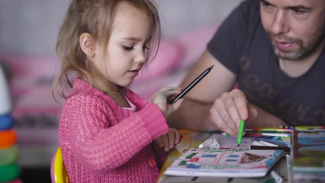 Little girl with blond hair, pulled back in a ponytail, sitting at the children's table and draws a colored felt-tip pens in the artistic album. Her dad sitting next and help to choose the pan