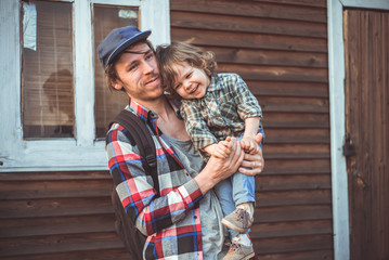 Папа и сын смеются в клетчатых рубашках на фоне деревянного дома