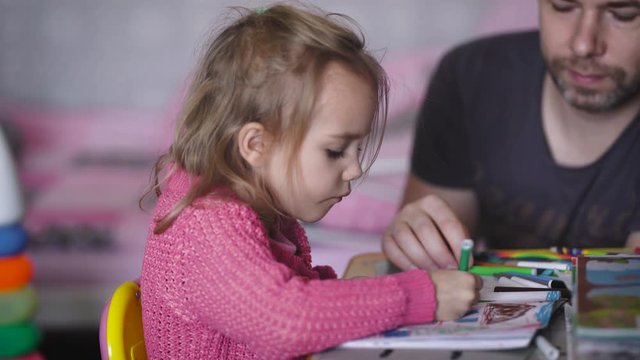 The baby with the father in children's to the room. The child with the man at a drawing table. The girl in a pink jacket paints a drawing album. The man has helped it to finish drawing a cloudlet.