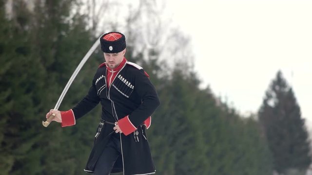 A young man in Cossack clothes swinging a sword in a winter landscape in the snow.