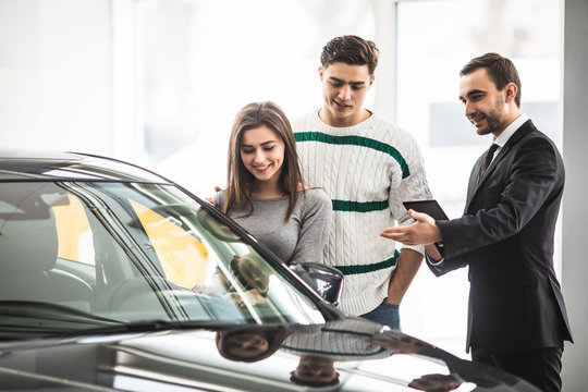 Beautiful Young Couple Choosing A Car At The Dealership Talking To The Salon Manager With Tablet In Hands