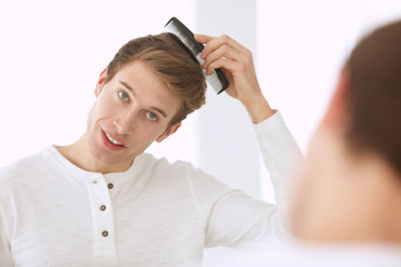 Handsome young man combing hair while standing in front of mirror