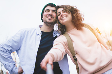Beautiful girl with a backpack and a guy in a light shirt taking selfie with selfie stick on a city street